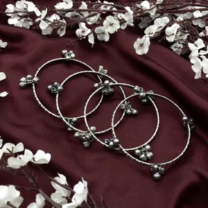 Puede incluir: Cuatro pulseras plateadas con adornos florales y de campanas se exhiben sobre una superficie de satén burdeos, acentuadas por ramas florales blancas. Las pulseras tienen un diseño texturizado y están adornadas con pequeños dijes colgantes.