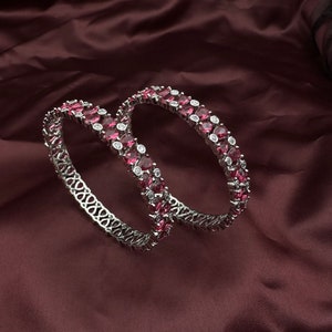 Pode incluir: Duas pulseiras prateadas com um padrão repetitivo de pedras preciosas ovais de cor rubi e pequenas pedras redondas e transparentes. As pulseiras são exibidas sobre um tecido bordô escuro.