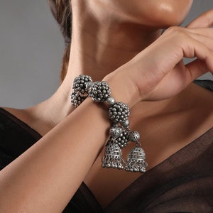 May include: A silver bracelet featuring a cluster of round beads and two dangling bells. The bracelet is crafted from metal and has a traditional design.