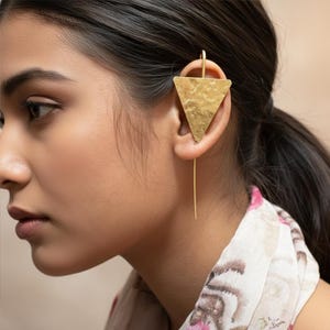 Op de afbeelding: Een goudkleurige oormanchet met een getextureerd driehoekig ontwerp. De oormanchet wordt op het oor gedragen en heeft een lang, dun, hangend element. Een uniek en modern accessoire.