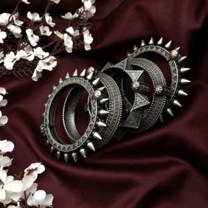 May include: Several silver-toned bangles with a spiked design are arranged on a deep red satin surface. The bangles have intricate patterns and are accented with small gold-colored details. White flowers are in the background.
