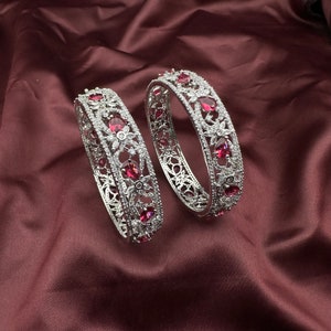 May include: Two silver bangles with intricate floral designs and pink gemstones. The bangles are on a burgundy fabric background.