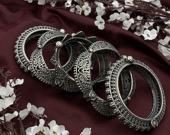 May include: A set of four silver-toned bangles with intricate floral designs. The bangles are stacked on top of each other and have a slightly curved shape.