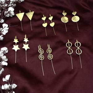 May include: A collection of gold-colored earrings in various designs, including triangles, hearts, stars, spirals, and figure-eight shapes. Each earring has a long, slender drop. The earrings are displayed on a dark burgundy fabric background.