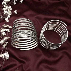 May include: Two silver arm cuffs. One cuff is a stack of curved bands with etched floral designs. The other cuff is a stack of plain curved bands. Both cuffs are on a burgundy background with white flowers.