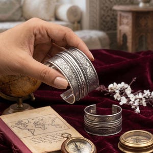 Puede incluir: Un brazalete de plata con intrincados patrones florales y geométricos se sostiene en una mano. El brazalete tiene una banda ancha con múltiples secciones texturizadas. Un segundo brazalete similar descansa sobre una superficie de terciopelo burdeos, junto con una brújula y un mapa antiguo.