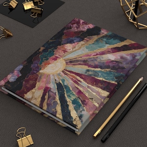 May include: A hardcover journal with a colorful abstract design featuring a sunburst pattern in shades of purple, teal, pink, and gold. The journal is closed, with a gold and black pencil resting beside it. Gold binder clips are also visible.
