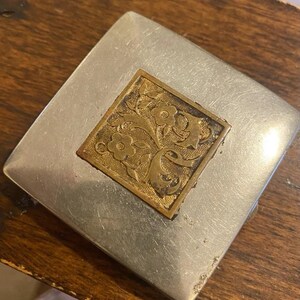 Op de afbeelding: Een zilverkleurig, vierkant compact poederdoosje met een decoratief, goudkleurig bloemmotief op de bovenkant. Het poederdoosje heeft een licht versleten uiterlijk, wat suggereert dat het vintage of antiek is.