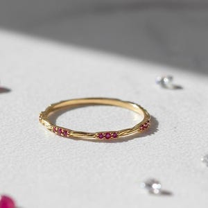May include: A gold ring with a textured band and small red gemstones. The ring is centered on a white surface, surrounded by scattered red and clear gemstones. The Nice Jewellery Shope logo is in the upper right corner.
