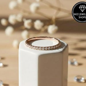 Puede incluir: Un anillo de oro rosa con una banda de pequeñas piedras preciosas transparentes. El anillo se exhibe sobre un soporte geométrico blanco, con un fondo borroso de flores blancas y otros anillos. La imagen incluye un logotipo que dice "Nice Jewellery Shope".