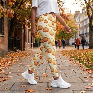 May include: Leggings featuring an all-over print of orange pumpkins on a cream-colored background. The leggings are paired with a plain white short-sleeved crop top and chunky white athletic sneakers, creating a casual and seasonal look.