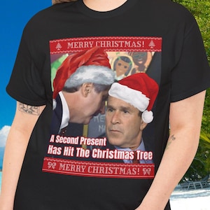 Puede incluir: Camiseta negra con un diseño navideño humorístico. El gráfico muestra a dos hombres con gorros de Papá Noel, con el texto "A Second Present Has Hit The Christmas Tree" y "MERRY CHRISTMAS!". La camiseta la lleva una persona frente a un fondo tropical.