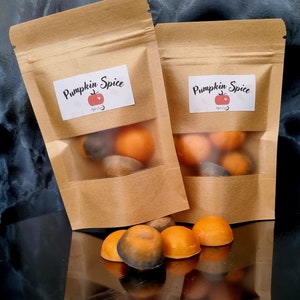 May include: Two brown paper bags with labels that say "Pumpkin Spice" and contain orange and brown wax melts. The bags are on a black surface with a reflection of the wax melts.