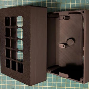 Op de afbeelding: Zwarte rechthoekige behuizing met een raster van vierkante openingen aan één kant en een apart deksel. De binnenkant van de behuizing is zichtbaar, met bevestigingskenmerken. Het product lijkt 3D-geprint te zijn.