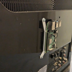 Könnte beinhalten: Eine kleine grüne Platine mit einer weißen Kunststoffhalterung, die an der Rückseite eines schwarzen Fernsehers befestigt ist. Die Platine hat mehrere Anschlüsse und Steckverbinder, darunter einen Ethernet-Anschluss, einen Micro-USB-Anschluss und einen HDMI-Anschluss.