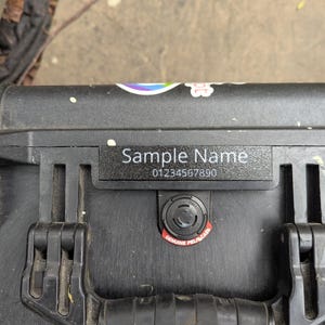 Può includere: Primo piano di una valigia nera con una targhetta con la scritta "Sample Name" e un numero di telefono. La valigia ha due chiusure nere e un adesivo rosso e bianco con la scritta "Genuine Pelican Case".