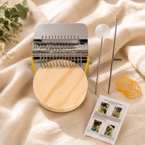 May include: A metal darning loom tool with a circular wooden base, a set of silver metal hooks, a long metal needle with a white spherical top, a thin metal hook tool, several yellow rubber bands, and a small instructional card with images.
