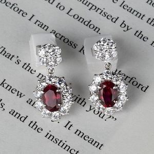 May include: A pair of silver drop earrings with a ruby-colored oval center stone, surrounded by a halo of small, sparkling white stones. The earrings are displayed on a book with printed text.