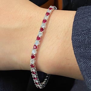 May include: A tennis bracelet featuring alternating red gemstones and clear, round-cut stones. The bracelet is set in a silver-colored metal and is displayed on a person's wrist, against a dark background.