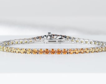 May include: A fine bracelet with a line of small, round gemstones in a colour gradient, shifting from pale yellow to a warm orange. The bracelet is set on a silver-toned metal band and is displayed on a white surface.