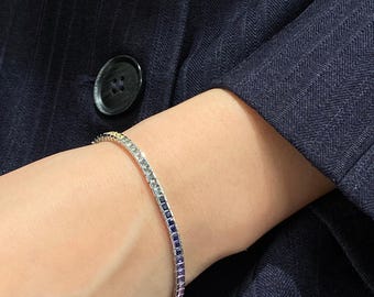 May include: A tennis bracelet featuring a continuous row of square-cut gemstones in a gradient colour scheme, transitioning from clear white to blue, purple, and pink. The stones are set in a metallic band that wraps around the wrist.