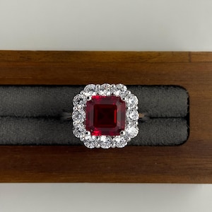 May include: A silver-toned ring featuring a large, square-cut deep red gemstone at the center. The central stone is surrounded by a halo of twelve small, round, clear white stones set in a metallic frame, creating a classic, symmetrical design.