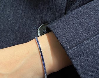 May include: A tennis bracelet featuring a continuous row of square-cut blue gemstones set in a metallic band. The stones transition in colour from a deep, saturated navy blue to a lighter, pale blue hue along the length of the bracelet.