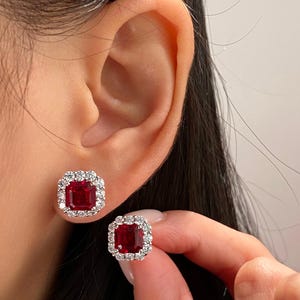 May include: A pair of stud earrings featuring a central emerald-cut deep red gemstone surrounded by a halo of small, round, clear white stones set in a metallic frame. One earring is positioned on the earlobe and the other is held by fingers.