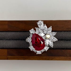Puede incluir: Un anillo de plata con una gran gema roja en forma de pera, rodeada de piedras blancas más pequeñas. El anillo tiene un diseño floral con piedras blancas y amarillas. El anillo se exhibe sobre una superficie de madera.