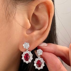 May include: A pair of silver-toned drop earrings featuring a central oval-cut red gemstone surrounded by a halo of small clear white stones. Above the main drop, a smaller flower-shaped cluster of clear white stones connects to the earring post.