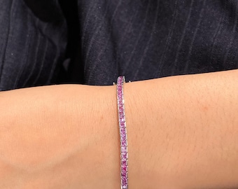 May include: A tennis bracelet featuring a continuous row of square-cut, vibrant pink gemstones set in a polished silver-toned metal band. The bracelet is secured with a clasp and sits firmly around the wrist, showcasing a uniform design.