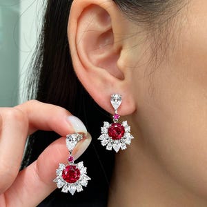May include: Dangling earrings featuring a large, round, red gemstone surrounded by smaller white gemstones in a floral design. A smaller red gemstone and a pear-shaped white gemstone are above the floral design. The earrings are set in a silver metal.