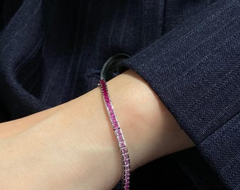 May include: A tennis bracelet featuring a row of square-cut stones that transition in colour from deep magenta to light pink. The stones are set in a metallic band, creating a gradient effect that wraps around the wrist in a continuous line.