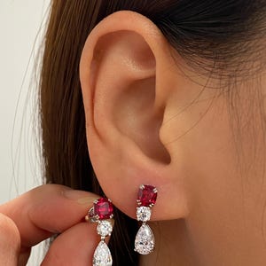 May include: A pair of drop earrings featuring a cushion-cut red gemstone at the top, followed by a small round clear gemstone, and finished with a pear-shaped clear gemstone at the bottom. The stones are set in a silver-toned metal setting.