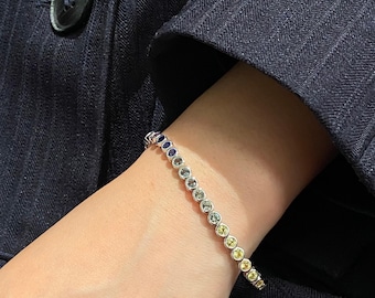 May include: A silver-toned tennis bracelet featuring a row of round, bezel-set gemstones in a gradient of colors, transitioning from deep blue at one end to light yellow and gold tones at the other, secured with a metallic link chain.