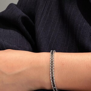 Op de afbeelding: Een zilverkleurige tennisarmband met een rij afwisselend vierkant geslepen donkerbruine stenen en kleine heldere witte steentjes in een metalen zetting, gedragen om de pols tegen een donkerblauwe stof met krijtstreep.