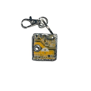 May include: A square circuit board keychain with a silver clasp and ring. The circuit board has yellow and brown detailing. The text "6BPW78" and "6BPW56" is printed on the board. A unique tech-themed accessory.
