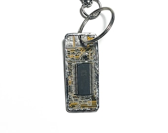 May include: A rectangular keychain featuring a circuit board design with a silver-colored metal clasp and key ring. The circuit board has a dark gray integrated circuit chip in the center. The keychain is suspended against a white background.