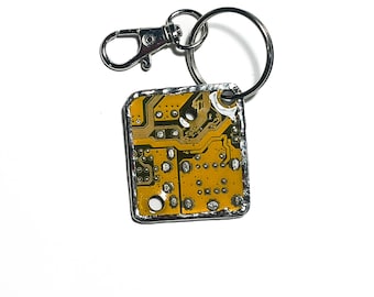 May include: A square keychain featuring a circuit board design. The yellow and green circuit board is encased in a silver-colored metal frame. The keychain includes a silver-colored split ring and a lobster clasp for easy attachment.