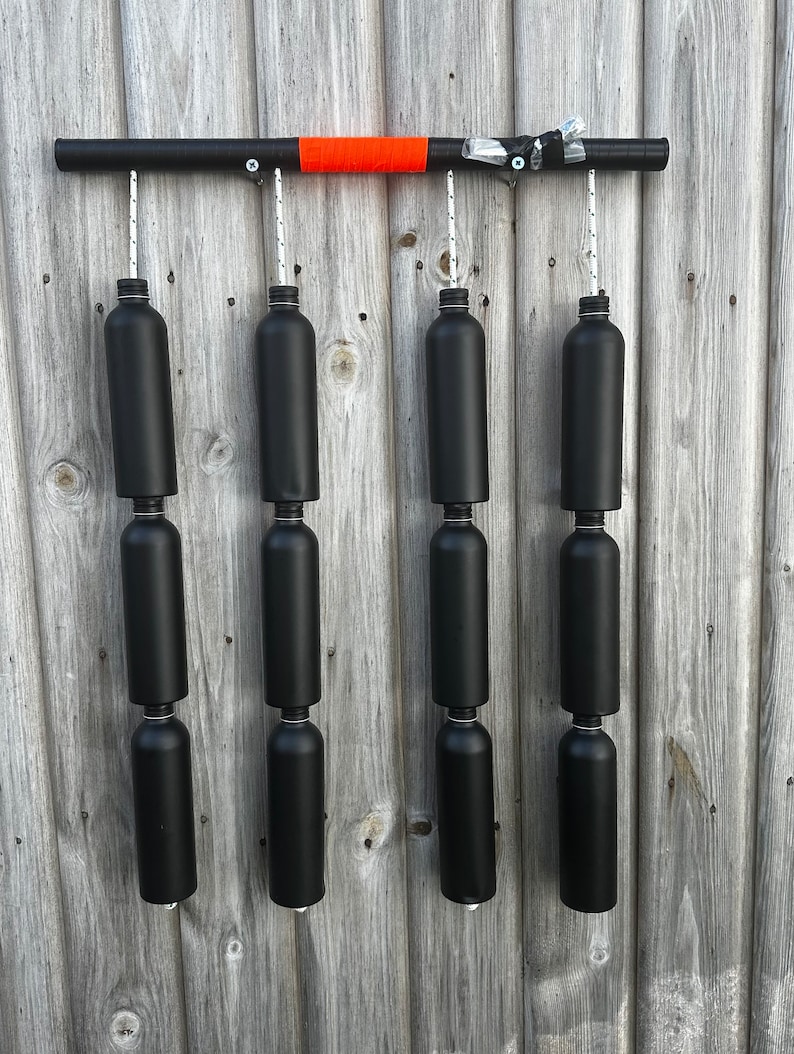 May include: A black metal bar with an orange stripe hangs from a wooden wall. Six black plastic bottles are suspended from the bar by ropes.
