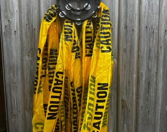 May include: A black tambourine with yellow caution tape streamers attached to it. The tambourine is hanging on a wooden wall.