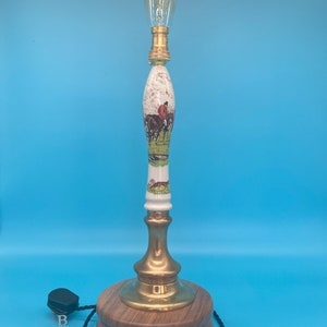 May include: A table lamp with a white ceramic base featuring a hunting scene. The base is topped with a brass finial and a clear glass bulb. The lamp sits on a wooden base.