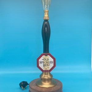 May include: A vintage-style lamp made from a repurposed beer pump handle. The lamp features a clear glass bulb and a wooden base. The beer pump handle has a red and white label that reads "Morland "Old Speckled Hen" Strong-Free Ale 5.1% Vol."
