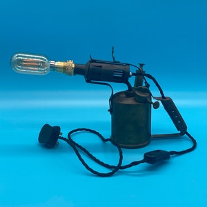 Może przedstawiać: Lampa w stylu vintage wykonana z mosiądzu w kształcie palnika z czarnym kablem i wtyczką. Lampa ma przezroczystą żarówkę i czarny metalowy klosz.