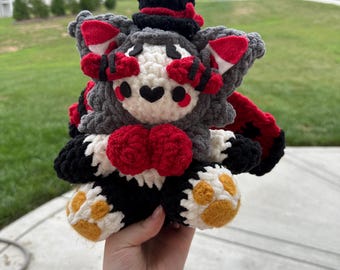 May include: A handmade crochet plush toy in the shape of a creature with a top hat. The toy features a gray body, red accents, and a black top hat. The paws are white with yellow paw pads. The toy is held in a hand.