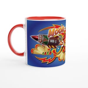 Puede incluir: Taza de cerámica blanca con asa e interior rojos. La taza presenta un diseño retro con un cohete y el texto "MOON" en una fuente estilizada. El cohete es marrón, rojo y verde, sobre un fondo azul.