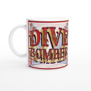 May include: White ceramic mug featuring the words "DIVE BOMBER" in red and gold lettering. The design includes star accents and a red border. The mug has a white handle.