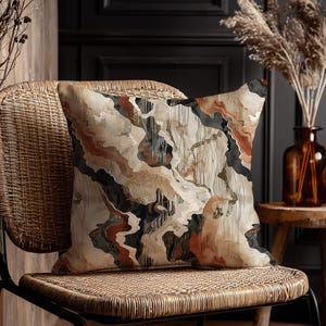 Puede incluir: Un cojín decorativo con un diseño abstracto en tonos beige, marrón, negro y óxido. El cojín está colocado sobre una silla de mimbre con estructura de metal negro. El diseño del cojín se asemeja a un patrón veteado o geológico.