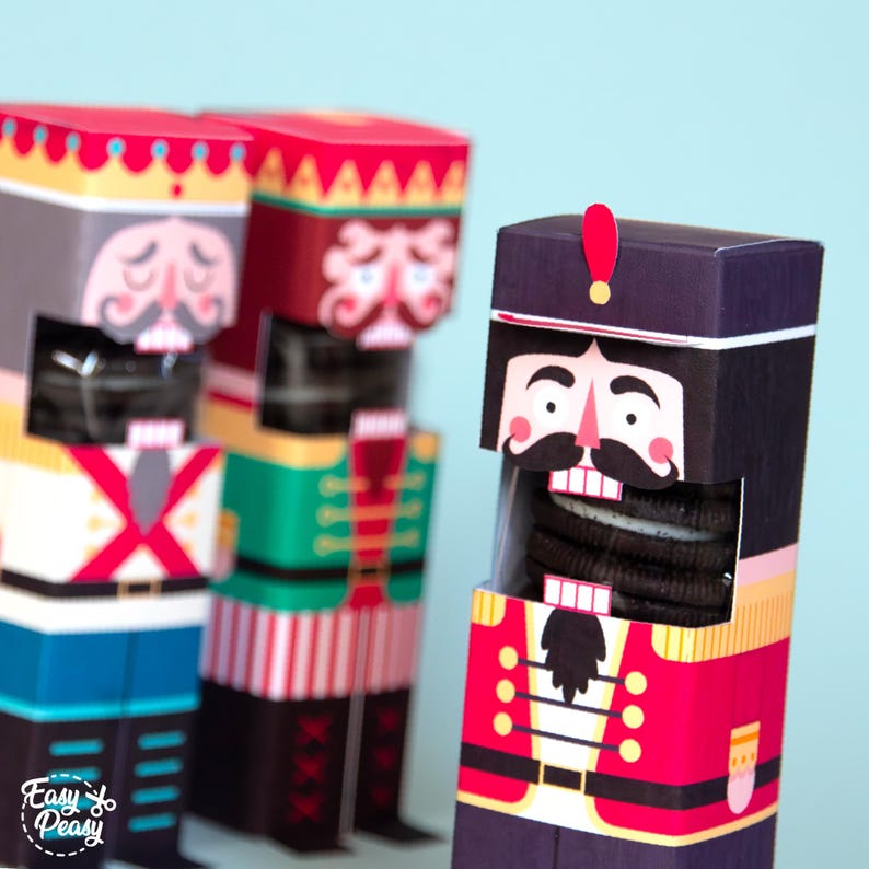 May include: Three nutcracker-themed cookie boxes. The boxes are colorful, with red, green, and blue accents. Each box has a different nutcracker face design. The boxes are filled with chocolate sandwich cookies.