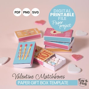 May include: A collection of Valentine's Day-themed matchboxes. The matchboxes are in various colors, including pink, blue, and yellow, with text that reads "We're a Perfect Match" and "You're Hot". The image also includes heart-shaped decorations.